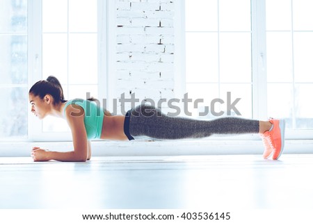 Perfect plank. Full-length side view of young beautiful woman in sportswear doing plank while standing in front of window at gym