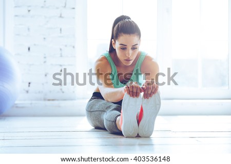 Dedicated to fitness. Front view of beautiful young woman in sportswear doing stretching while sitting on the floor in front of window at gym