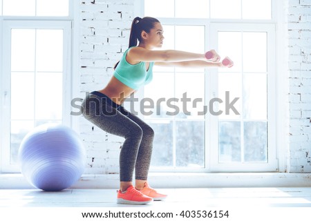 Deep squat. Side view of young beautiful woman in sportswear doing squat and holding dumbbells while standing in front of window at gym
