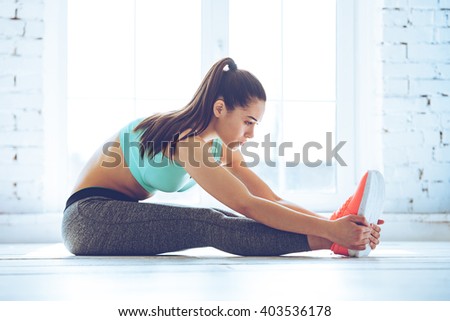Stretching those muscles before workout. Side view of beautiful young woman in sportswear doing stretching while sitting on the floor in front of window at gym