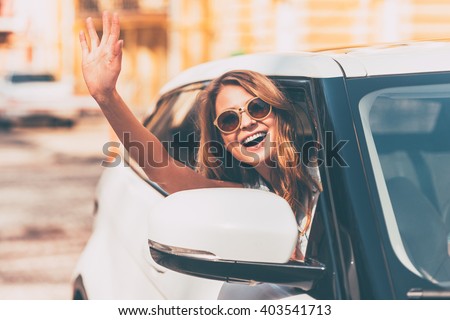 Hello! Beautiful young cheerful women looking at camera with smile and waving while sitting in her car
