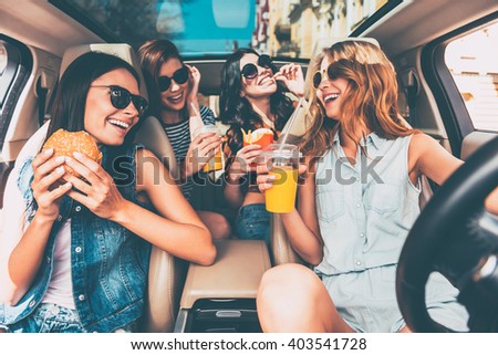 Enjoying their lunch in the car. Four beautiful young cheerful women looking at each other with smile and eating take out food while sitting in car