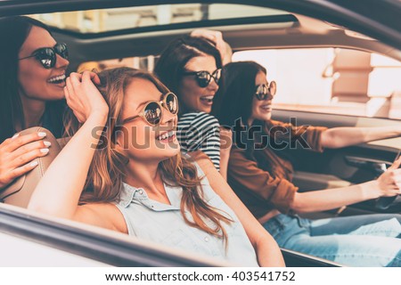 On the road together. Side view of four beautiful young cheerful women looking away with smile while sitting in car