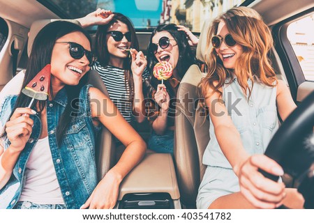 Great start of their journey. Four beautiful young cheerful women looking at each other with smile and holding lollipops while sitting in car