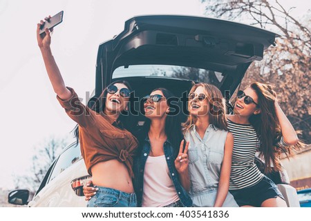 This is why we take road trips! Four beautiful young cheerful women making selfie with smile while standing near car trunk