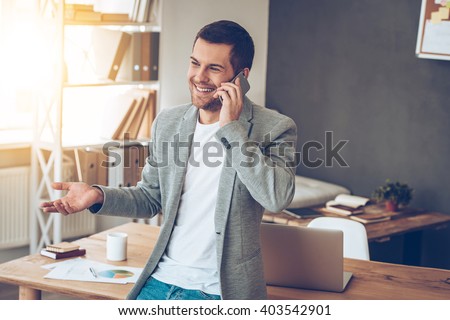 See you in my office! Handsome young man talking on mobile phone with smile while leaning to the table at his office