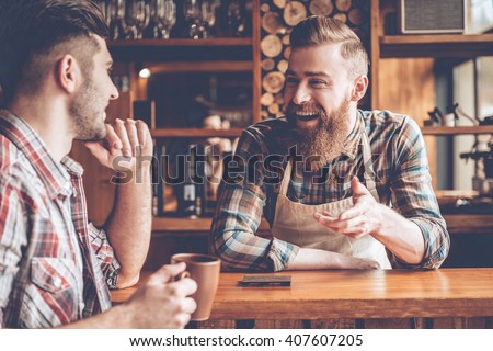 Sharing good news. Barista and his customer discussing something with smile while sitting at bar counter at cafe