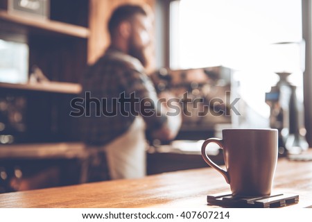 Morning coffee. Focused picture of coffee cup standing at bar counter with young bearded man in apron making coffee in background