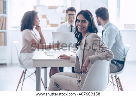 Happy to be in team. Beautiful cheerful woman looking at camera with smile while sitting at the office table with her coworkers