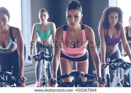 Up to speed with their fitness goals. Young beautiful women with perfect bodies in sportswear looking at camera while cycling at gym