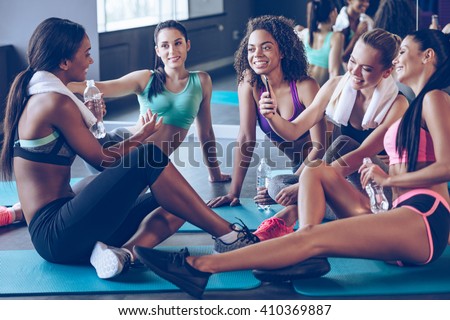 Meet up on the mat. Beautiful young women in sportswear discussing something with smile and using smartphone while sitting on exercise mat at gym