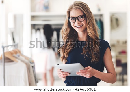 Modern businesswoman. Beautiful young woman holding digital tablet and looking at camera with smile while standing at the clothing store