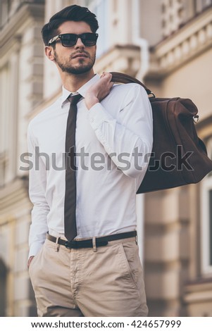 Trendy and handsome. Low angle view of handsome young man in sunglasses holding leather bag and looking away while walking outdoors