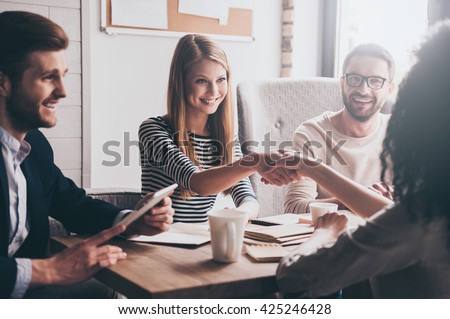 We have a deal! Two beautiful women shaking hands and looking at each other with smile while sitting at the business meeting with their coworkers