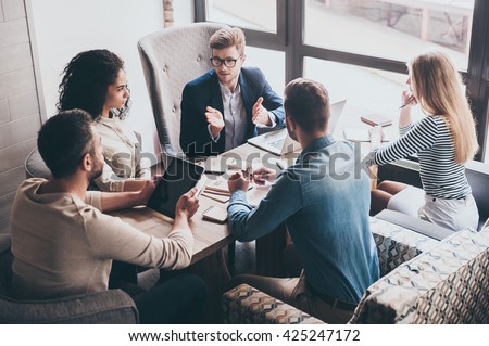 Combining their expertise. Young handsome man in glasses gesturing and discussing something with his coworkers while sitting at the office table