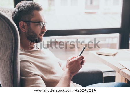 Business message. Side view of handsome man using his smartphone while sitting in chair in front of window