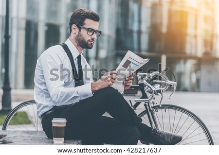 Reading fresh news. Side view of young businessman reading newspaper while sitting near his bicycle with office building in the background