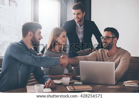 We have a deal! Two men shaking hands and looking at each other with smile while sitting at the business meeting with their coworkers