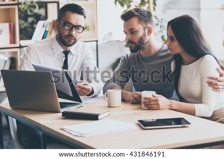 I need your signature here. Confident young man in shirt and tie holding some document and pointing it while sitting together with young couple at the desk in office