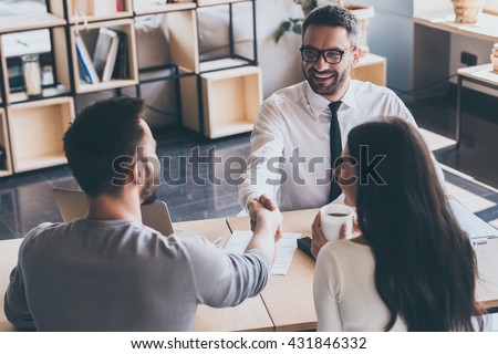 Sealing a deal. Top view of two men sitting at the desk and shaking hands while young woman looking at them and smiling