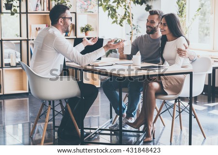 Good news for you! Cheerful young couple bonding to each other and listening to their financial advisor sitting at the desk in front of them