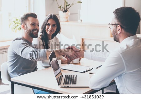 Good deal! Cheerful young man bonding to his wife while shaking hand to man sitting in front of him at the desk