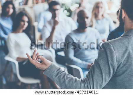 Business coach. Rear view of man gesturing with hand while standing against defocused group of people sitting at the chairs in front of him