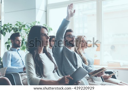 I have a question! Group of young people sitting on conference together while one man raising his hand