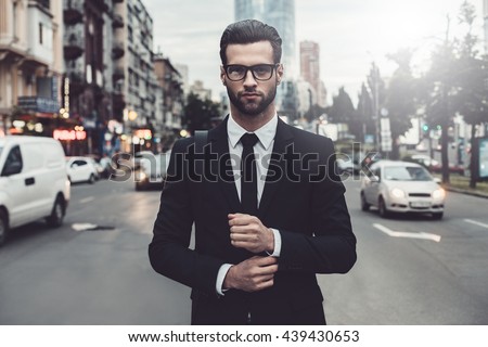 Confident businessman. Confident young man in full suit adjusting his sleeve and looking away while standing outdoors with cityscape in the background