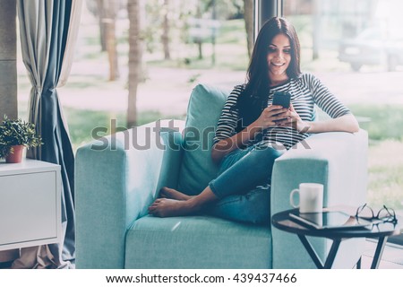 Lovely message from him. Beautiful young smiling woman looking at her smart phone while sitting in a big comfortable chair at home