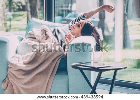 Enjoying beautiful morning. Beautiful young woman stretching out hands and smiling while lying in a big comfortable chair at home