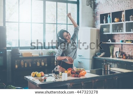 Cooking with passion. Beautiful young mixed race woman in headphones cooking salad and dancing while standing in kitchen at home