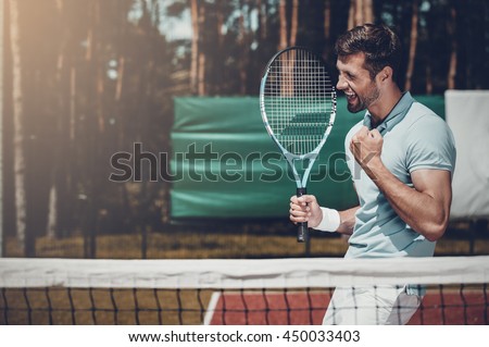 Happy winner. Side view of happy young man in polo shirt holding tennis racket and gesturing while standing on tennis court