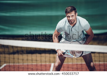 Tennis player. Handsome young man in polo shirt holding tennis racket and smiling while standing on tennis court