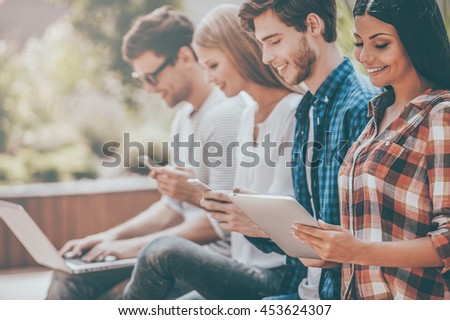 Living in digital age. Group of happy young people holding different digital devices and smiling while sitting in a row outdoors