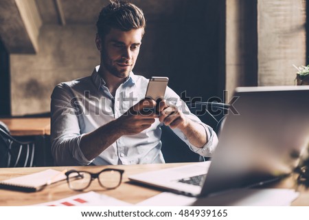 Texting to colleague. Confident young man in smart casual wear holding smart phone and looking at it while sitting at his working place in office