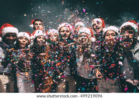 Colorful fun. Group of beautiful young people in Santa hats blowing colorful confetti and looking happy