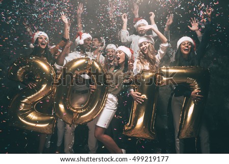 Happy New Year to you! Group of cheerful young people in Santa hats carrying gold colored numbers and throwing confetti