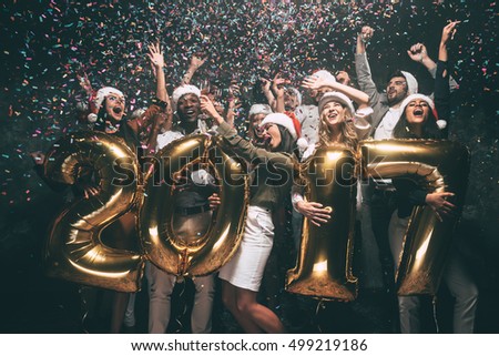 Celebrating New Year. Group of cheerful young people in Santa hats carrying gold colored numbers and throwing confetti
