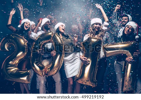 Wishing a happy New Year. Group of cheerful young people in Santa hats carrying gold colored numbers and throwing confetti