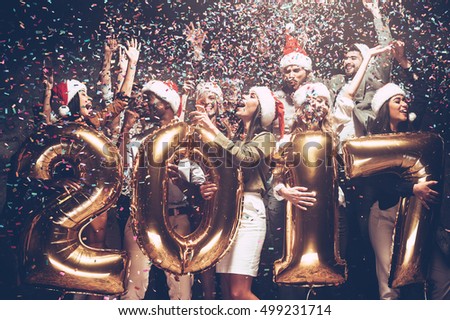 Happy New Year! Group of cheerful young people in Santa hats carrying gold colored numbers and throwing confetti