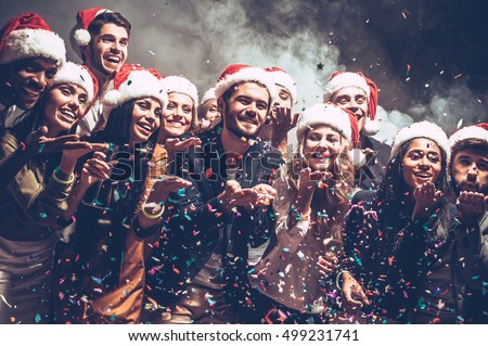 Little Christmas magic. Group of beautiful young people in Santa hats blowing colorful confetti and looking happy