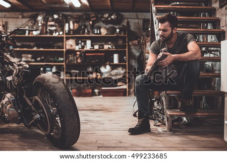 Confident mechanic. Confident young man holding rag and looking at motorcycle while sitting near it in repair shop