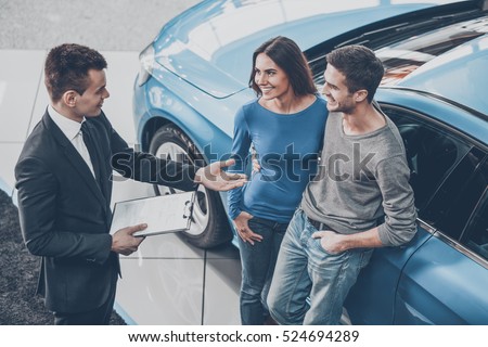 Buying their first car together. High angle view of young car salesman standing at the dealership telling about the features of the car to the customers