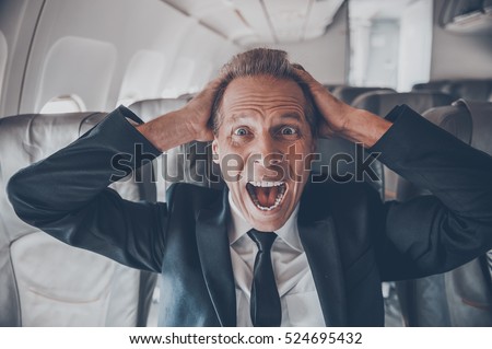 Fear of flight. Shocked mature businessman touching his head with hands and looking at camera while sitting at his seat in airplane