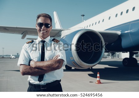 Confident pilot. Confident male pilot in uniform keeping arms crossed and smiling with airplane in the background