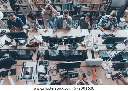 Busy working in office. Top view of group of young business people in smart casual wear working together while sitting at the large office desk