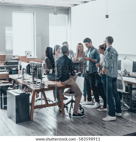 Working as one team. Full length of young modern people in smart casual wear having a meeting while standing behind the glass wall in the creative office