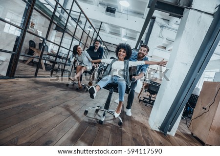 Work hard play hard! Four young cheerful business people in smart casual wear having fun while racing on office chairs and smiling