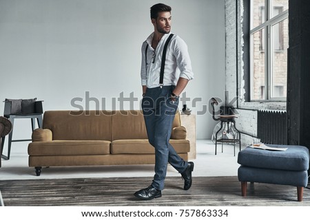 Time to go. Full length of handsome young man in white shirt and suspenders looking away and keeping hands in pockets while walking through the room